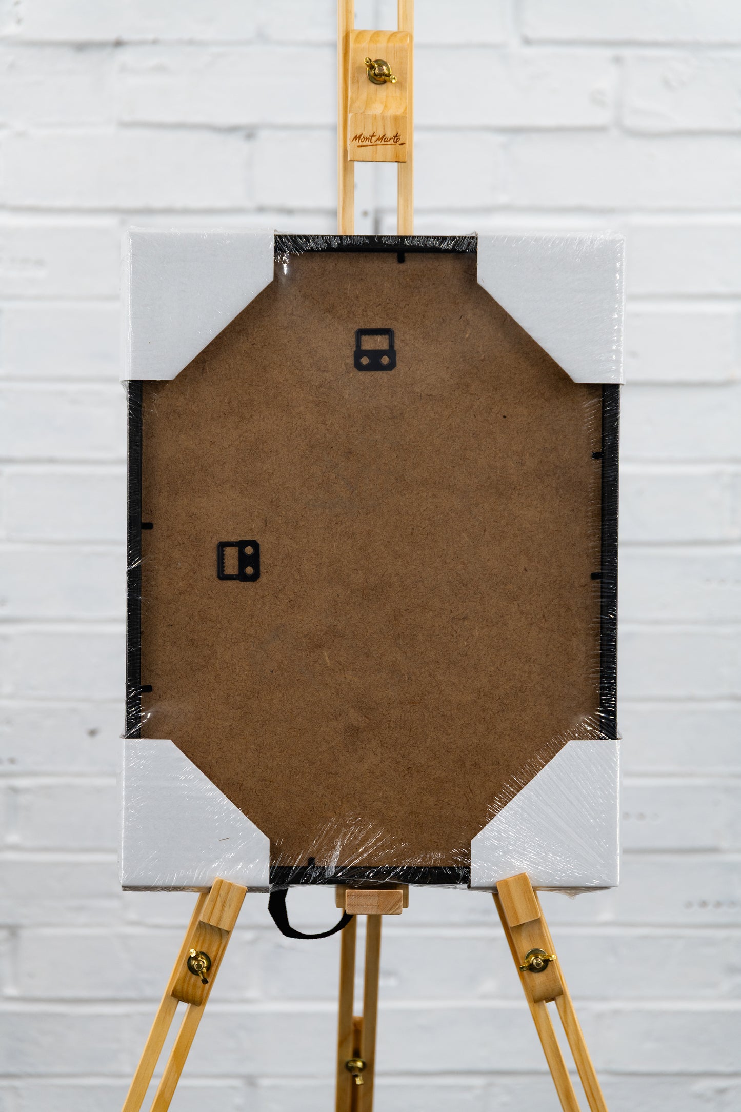 Rear of framed aviation print on an easel with MDF back panel, shrink wrap, and protective corner guards against a white brick wall.