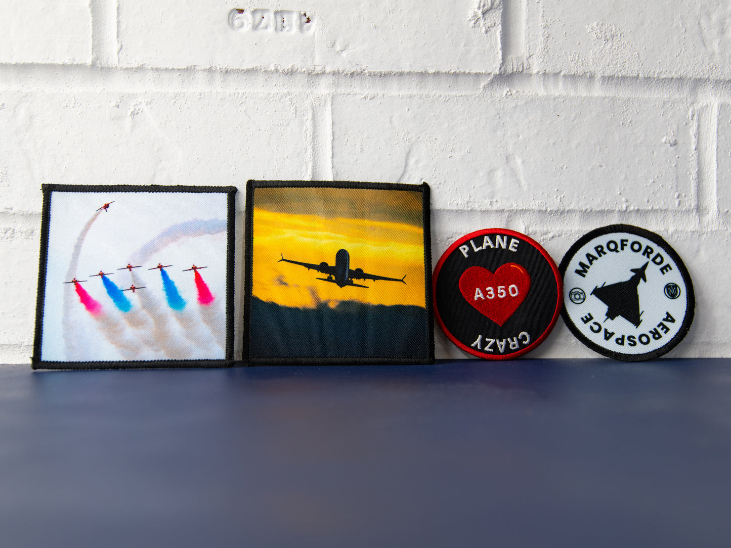 All four patches in a row against a white wall: Red Arrows photo art, sunset airliner photo art, Plane Crazy heart, Marqforde crest.