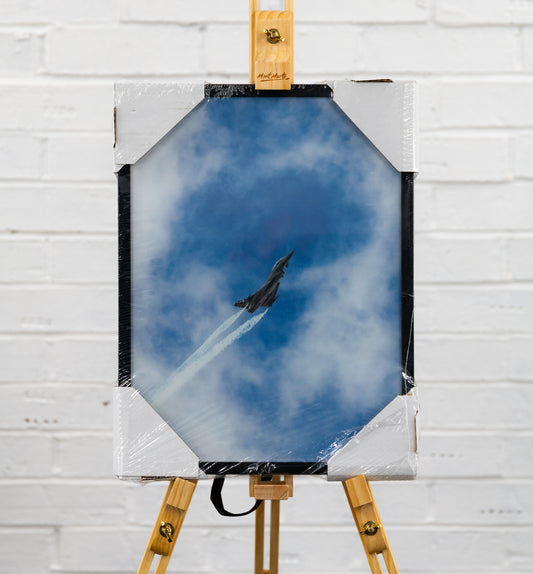 Front-on view of the framed print in factory wrap, showing the jet spearing up through blue sky and cloud