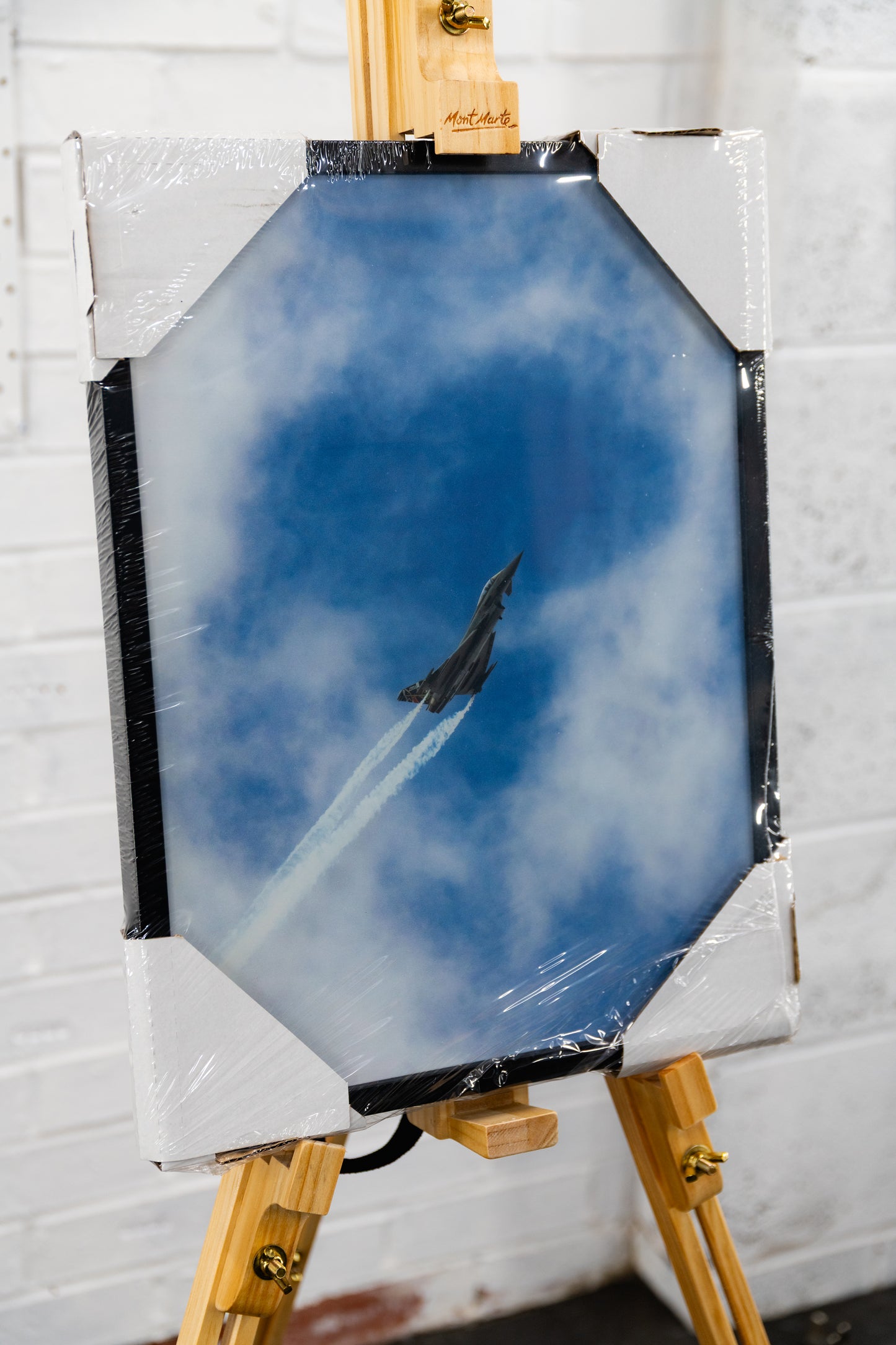 Three-quarter left view of framed Typhoon jet print—aircraft climbing with twin vapour trails through cloud—still in shrink wrap with corner protectors
