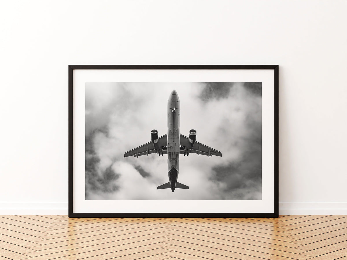 Airbus A320 passing directly overhead against bright cloud, black and white, aviation photo wall art print in a black frame